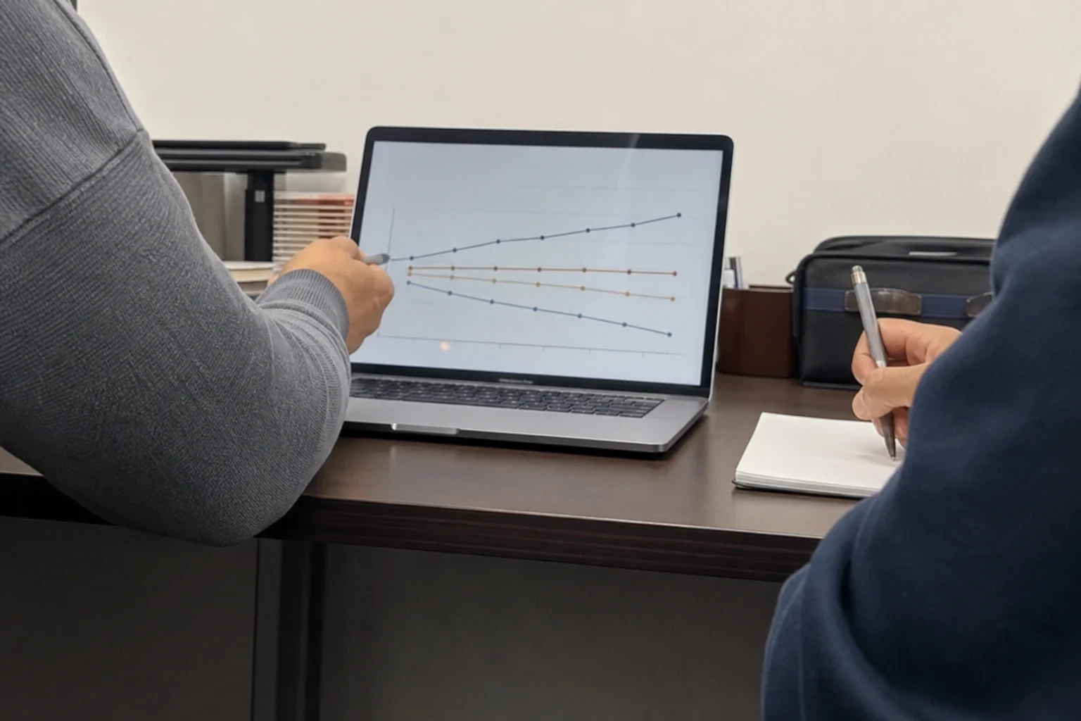 Person in a gray sweater points a pen at line graphs on a laptop while another person takes notes on a notepad at an office desk