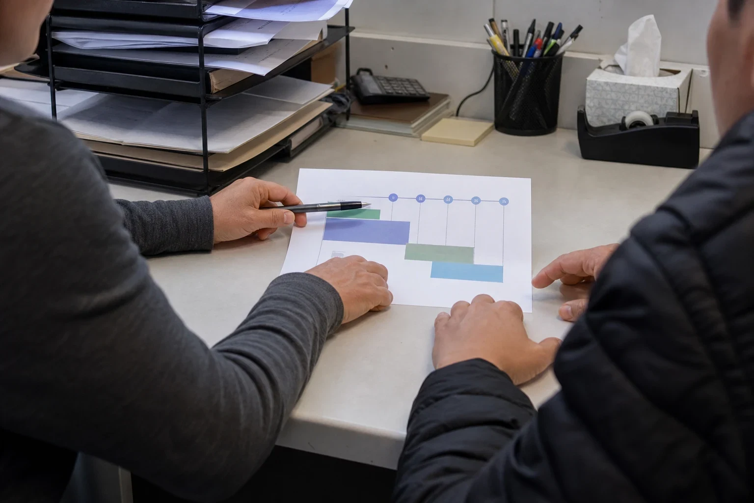 Two people at a cluttered office desk, one pointing a pen at a printed bar chart beside paper trays, a calculator, a pen cup and a tissue box